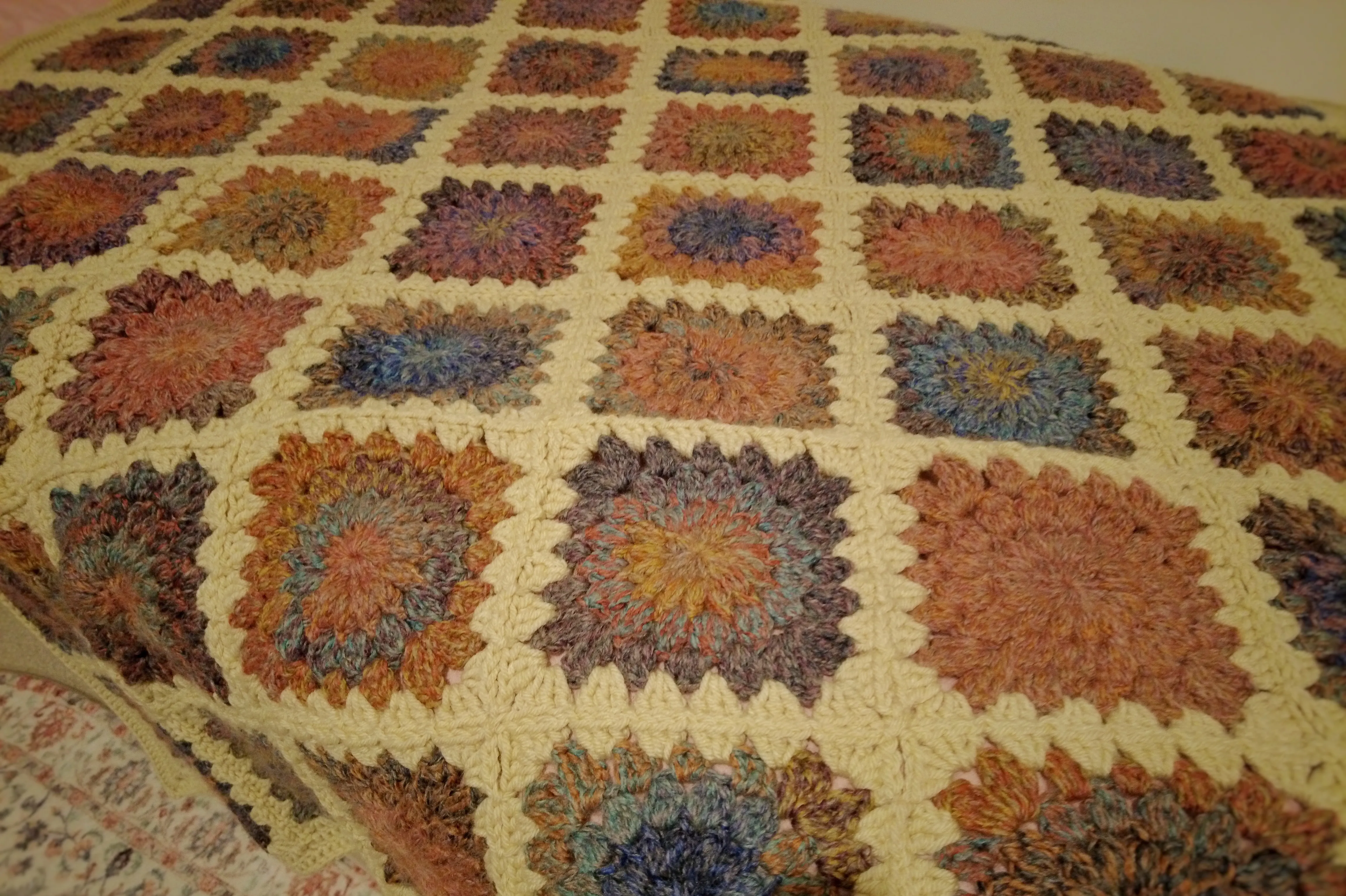 A crochet blanket spread out on a bed. It's made of granny squares in marbled rainbow hues, and they're connected by cream yarn stitches.